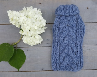 May include: A blue knitted sweater for a small dog, with a cable knit pattern. The sweater is laying on a light gray wooden surface.
