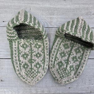 Puede incluir: Un par de zapatillas tejidas a mano con un dise&ntilde;o bicolor en verde salvia y beige avena. Las zapatillas presentan un patr&oacute;n geom&eacute;trico de rombos repetido en la parte superior, complementado con detalles de rayas verticales en el borde.