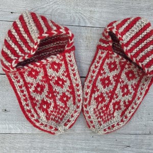Puede incluir: Un par de zapatillas tejidas a mano con un dise&ntilde;o bicolor en rojo y beige. Las zapatillas presentan un patr&oacute;n floral geom&eacute;trico repetitivo en la parte superior y un dise&ntilde;o de rayas en el pu&ntilde;o, confeccionadas con hilo grueso y texturizado.