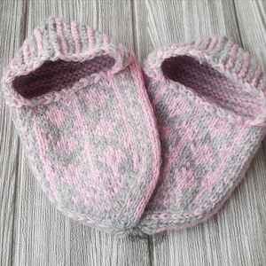 Puede incluir: Un par de zapatillas tejidas a mano con un patr&oacute;n de dos tonos en hilo rosa suave y gris claro. Las zapatillas tienen una abertura de pu&ntilde;o acanalado y un dise&ntilde;o geom&eacute;trico decorativo en la parte superior, con una textura de punto visible.
