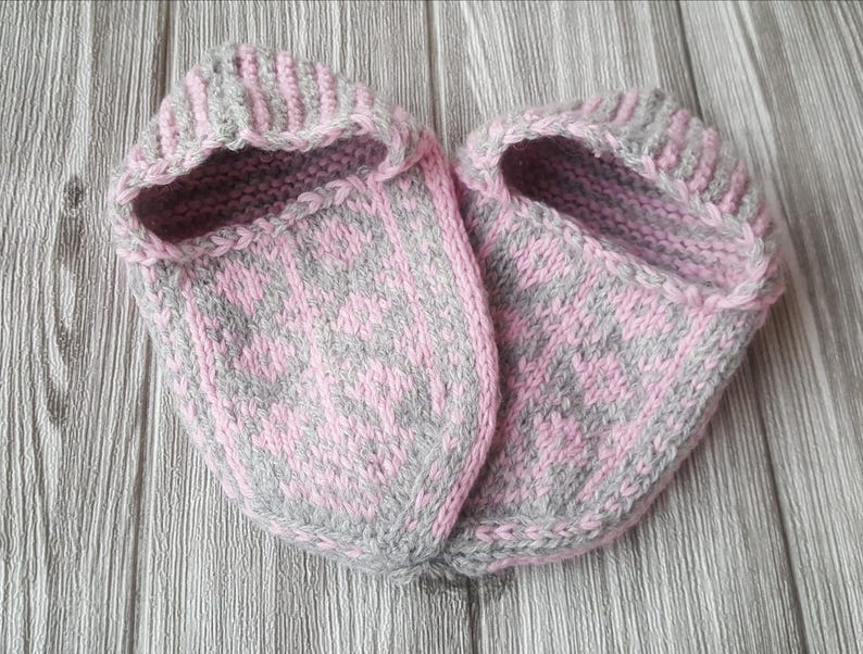 Puede incluir: Un par de zapatillas tejidas a mano con un patr&oacute;n de dos tonos en hilo rosa suave y gris claro. Las zapatillas tienen una abertura de pu&ntilde;o acanalado y un dise&ntilde;o geom&eacute;trico decorativo en la parte superior, con una textura de punto visible.