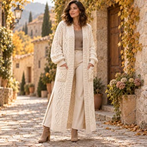 Könnte beinhalten: Eine lange, cremefarbene Strickjacke mit Zopfmuster, offener Vorderseite, langen Ärmeln und zwei aufgesetzten Taschen. Das Kleidungsstück wird über einem schlichten beigen Oberteil und einer hellen Hose getragen, kombiniert mit beigen Stiefeletten.