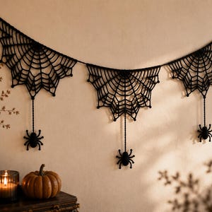 May include: A black crocheted garland featuring three interconnected spider web motifs. Each web section is suspended from a horizontal cord and features a small, dangling black spider attached by a thin, vertical crocheted thread.