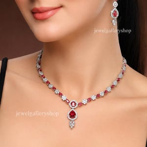 May include: A silver-toned necklace and matching drop earring set featuring oval-cut red stones surrounded by small clear white stones. The necklace has a central pendant with a pear-shaped red stone and a small leaf-like accent at the bottom.