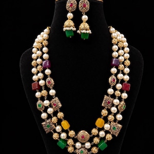 May include: A three-strand necklace and matching jhumka earrings set featuring white pearls, gold-toned filigree beads, and square-cut stones in red, green, and yellow. The earrings have a pearl stud and a bell-shaped drop with a green stone.