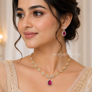 Puede incluir: Un conjunto de collar y pendientes colgantes en tono dorado con piedras rosas en forma de lágrima rodeadas de pequeños cristales transparentes. La cadena del collar presenta eslabones en forma de ala con cristales y un dije central.