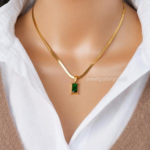 Op de afbeelding: Een goudkleurige visgraatketting met een rechthoekige smaragdgroene hanger. De hanger is gezet in een goudkleurig metalen frame. De tekst jewelgalleryshop is zichtbaar aan de rechterkant van de ketting.