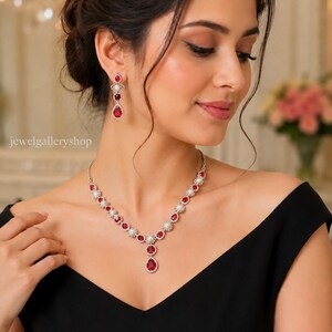 Può includere: Un set di collana e orecchini pendenti color argento con pietre rosse a forma di goccia alternate a perle bianche rotonde. La collana presenta un ciondolo centrale e gli orecchini riprendono il design con una pietra rossa più piccola sopra.