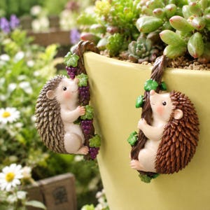 May include: Two small hedgehog figurines designed to hang on the rim of a planter. Each hedgehog is climbing a brown textured vine adorned with green leaves and clusters of purple grapes. The hedgehogs have light-colored faces and brown spines.