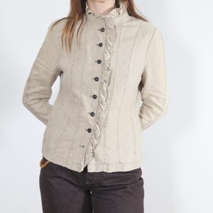 May include: Beige jacket with a high ruffled collar and a row of black buttons down the front. The jacket has a fitted silhouette and is paired with dark brown pants. The jacket's seams are accented with darker stitching.