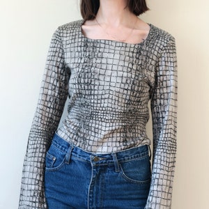 May include: A long-sleeved blouse with a square neckline and a crocodile print pattern. The blouse is beige with black accents. The model is wearing blue jeans.