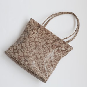 Könnte beinhalten: Eine beige Tasche mit Schlangenhautmuster und doppelten Henkeln.