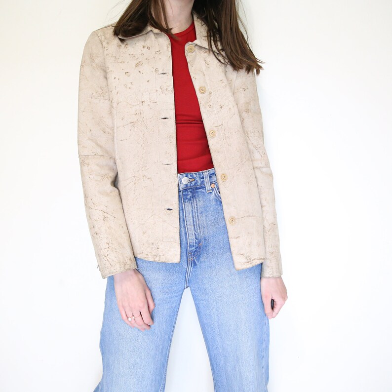 K&ouml;nnte beinhalten: Eine beige Wildlederjacke mit Knopfleiste und einem strukturierten Muster. Die Jacke wird &uuml;ber einem roten Shirt und einer blauen Jeans getragen.