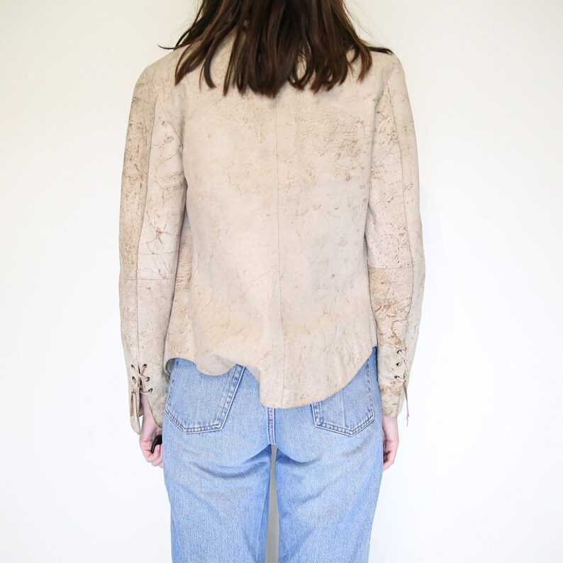 K&ouml;nnte beinhalten: Eine beige Wildlederjacke mit Used-Look. Die Jacke hat einen Schn&uuml;rdetail an den &Auml;rmeln und einen leicht verk&uuml;rzten Saum.