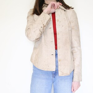 K&ouml;nnte beinhalten: Eine beige Wildlederjacke mit Kn&ouml;pfen und langen &Auml;rmeln. Die Jacke hat eine strukturierte Oberfl&auml;che und eine leicht verk&uuml;rzte Passform.