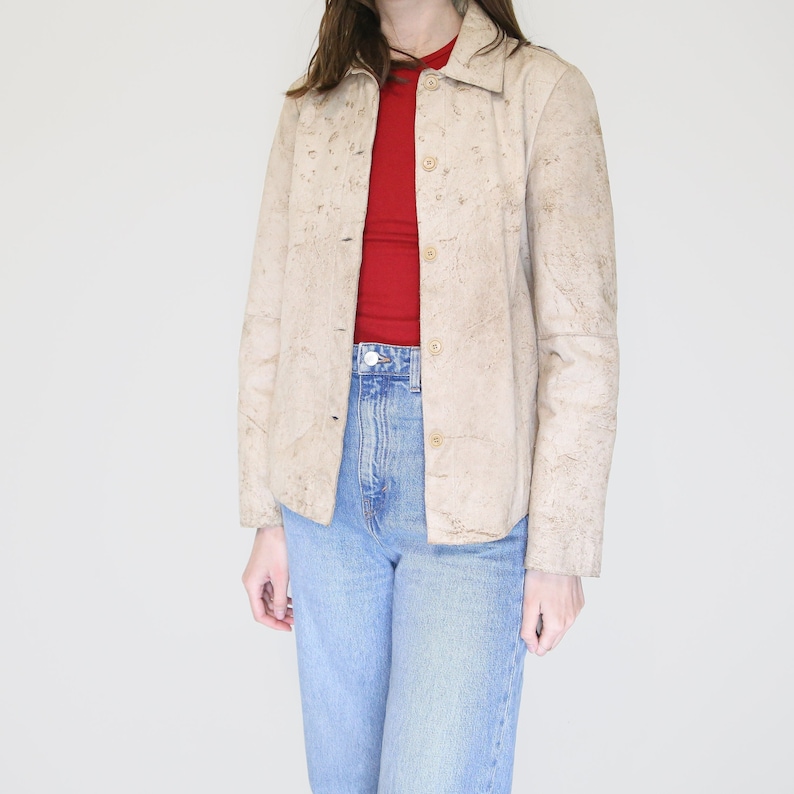 K&ouml;nnte beinhalten: Eine beige Wildlederjacke mit Knopfleiste und langen &Auml;rmeln. Die Jacke hat eine strukturierte, leicht abgenutzte Optik.