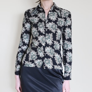 May include: A black button-down shirt with a floral pattern in white, green, and beige. The long-sleeved shirt has a pointed collar and is buttoned up. The floral design is repeated across the fabric.