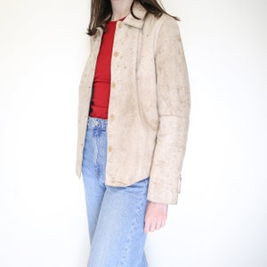 K&ouml;nnte beinhalten: Eine beige Wildlederjacke mit Knopfleiste und langen &Auml;rmeln. Die Jacke hat einen leicht abgenutzten Look.