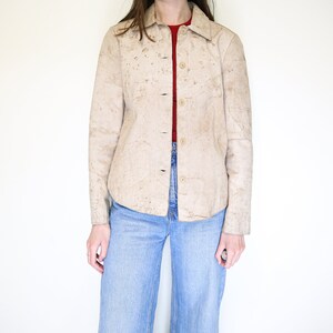 K&ouml;nnte beinhalten: Eine beige Wildlederjacke mit Kn&ouml;pfen und langen &Auml;rmeln. Die Jacke hat ein strukturiertes, gesprenkeltes Muster.