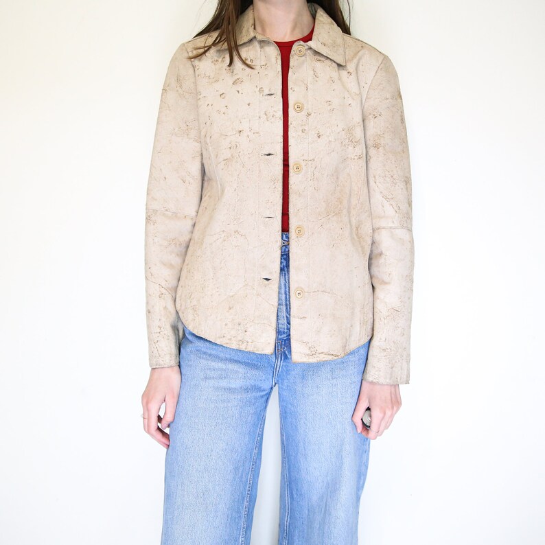 K&ouml;nnte beinhalten: Eine beige Wildlederjacke mit Kn&ouml;pfen und langen &Auml;rmeln. Die Jacke hat ein strukturiertes, gesprenkeltes Muster.