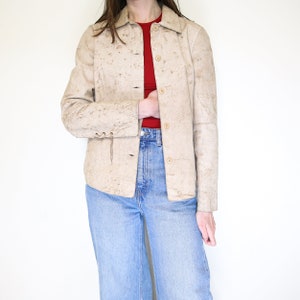 K&ouml;nnte beinhalten: Eine beige Lederjacke mit Knopfleiste und langen &Auml;rmeln. Die Jacke hat eine strukturierte, genarbte Oberfl&auml;che und eine leicht verk&uuml;rzte L&auml;nge.