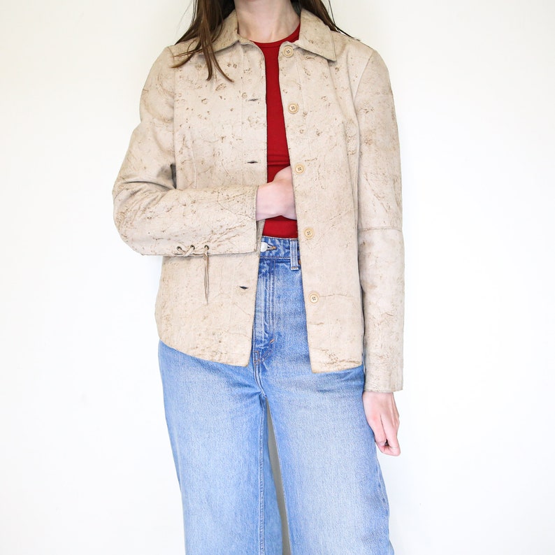 K&ouml;nnte beinhalten: Eine beige Lederjacke mit Knopfleiste und langen &Auml;rmeln. Die Jacke hat eine strukturierte, genarbte Oberfl&auml;che und eine leicht verk&uuml;rzte L&auml;nge.