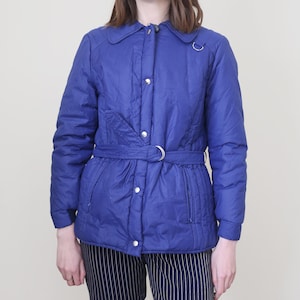 Könnte beinhalten: Eine blaue gesteppte Jacke mit Druckknopfverschluss, einem Gürtel und zwei Taschen. Die Jacke hat einen Kragen und lange Ärmel.