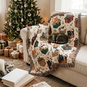 May include: A cream-coloured woven throw blanket featuring a repeating pattern of cats sitting inside gift boxes, with a tabby cat resting on top. The blanket has fringed edges and displays various cat colours including black, orange, and white.
