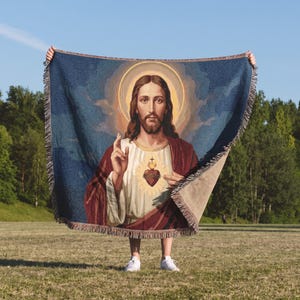 May include: A woven throw blanket featuring a depiction of the Sacred Heart of Jesus. The design shows Jesus with a halo, a red robe over a white tunic, and a heart with a cross. The blanket has a fringed edge and a dark blue background.