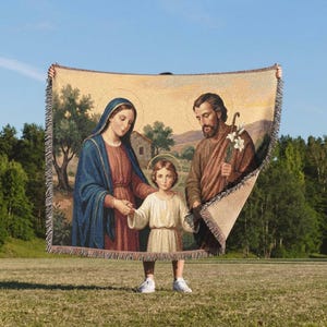 May include: A woven tapestry throw blanket featuring a depiction of the Holy Family. The image shows a woman in a blue and red robe, a man in brown robes holding a lily, and a child in a white tunic, all with halos against a landscape.