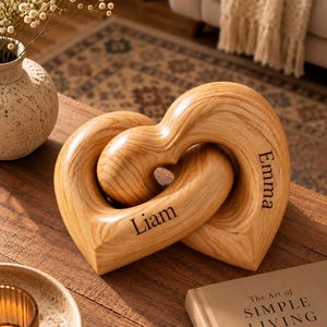 May include: A decorative wooden sculpture consisting of two interlocking heart shapes with a natural wood grain finish. The name Liam is engraved on the lower heart, and the name Emma is engraved on the upper heart in a dark, clear serif font.