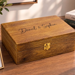 May include: A rectangular wooden keepsake box with a dark brown wood grain finish. The lid features the engraved text Daniel & Sophia in a cursive script. A gold-coloured metal latch is centred on the front edge of the box to secure the lid.