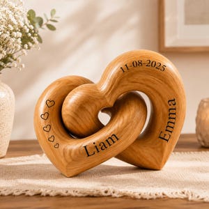 Puede incluir: Una escultura de madera de color marrón claro que consta de dos formas de corazón entrelazadas. La madera presenta una veta visible. En la superficie están grabados los nombres Liam y Emma, la fecha 11-08-2025 y cinco corazones pequeños.