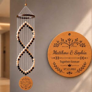 Può includere: Una giostrina sospesa con una struttura a forma di infinito composta da perline alternate nere e color crema appese a una cornice circolare in legno, accompagnata da una targa rotonda in legno incisa con Matthew & Sophia, Together Forever, 07.12.2024.