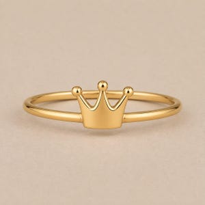 Peut inclure: Une bague en métal doré poli ornée d'un motif de couronne central. L'anneau est fin et circulaire, tandis que l'élément en forme de couronne présente trois pointes distinctes, chacune surmontée d'un petit détail sphérique, créant un design simple.
