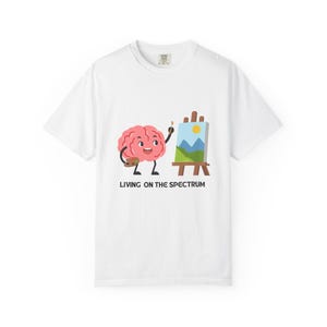 Op de afbeelding: Een wit t-shirt met een cartoonillustratie van een roze breinfiguur die een penseel en een palet vasthoudt en een landschap op een ezel schildert. Onder de afbeelding staat de tekst LIVING ON THE SPECTRUM in zwarte hoofdletters.