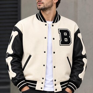 Puede incluir: Una chaqueta universitaria de color crema con mangas negras que presentan aplicaciones blancas en forma de hueso. La chaqueta tiene cuello, puños y dobladillo acanalados en negro con rayas blancas, un parche con la letra B y cierre de botones.