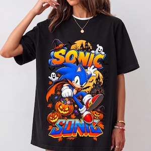 Könnte beinhalten: Ein schwarzes T-Shirt mit einem Grafikdruck von Sonic the Hedgehog in einem Vampirumhang, der einen mit Süßigkeiten gefüllten Kürbiseimer hält. Das Design zeigt den Schriftzug "SONIC" in blauen und orangen Buchstaben, Geister und ein Spukhaus.