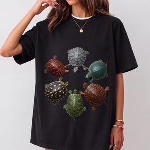 Peut inclure: Un t-shirt noir à manches courtes orné d'un imprimé graphique circulaire représentant cinq tortues différentes. Les tortues sont représentées dans diverses couleurs, notamment des tons de brun, jaune, vert et gris, disposées en cercle.