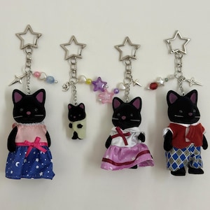 Op de afbeelding: Vier zwarte kattenfiguurtjes bevestigd aan zilveren stervormige sleutelhangers. De katten dragen diverse outfits, waaronder een blauwe jurk met sterrenprint, een wit met paarse rok en een rood vest met een blauwe broek met ruitjespatroon.