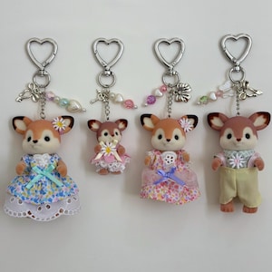 Op de afbeelding: Vier sleutelhangers met gevlokte hertenfiguren bevestigd aan zilverkleurige hartvormige sluitingen. Elk hert draagt stoffen kleding met bloemen- of effen patronen, geaccentueerd door kleine kralen, bedels en miniatuurbloemen aan de kettingen.