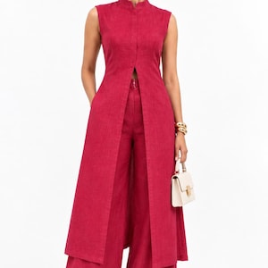 May include: A sleeveless magenta tunic with a high collar and a long front slit, paired with matching wide-leg trousers. The outfit features a textured fabric, a clean silhouette, and is accessorised with a small cream handbag and gold jewellery.