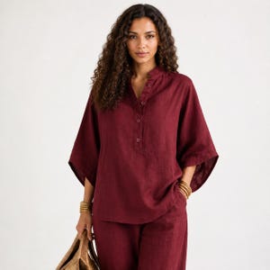 May include: A deep burgundy linen shirt featuring a mandarin collar, a partial button-down front with four visible buttons, and wide, elbow-length sleeves. The shirt has a relaxed, loose-fitting silhouette and a textured fabric appearance.