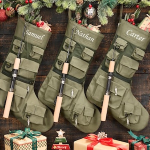 Puede incluir: Tres calcetines navideños de color verde oliva con múltiples bolsillos utilitarios, paneles de malla y una caña de pescar sujeta en la parte frontal. Cada calcetín está personalizado con nombres bordados en blanco: Samuel, Nathan y Carter.