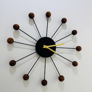 May include: A sunburst wall clock featuring a central black circular face with gold-colored hour and minute hands. Twelve thin black metal spokes radiate from the center, each tipped with a polished dark brown wooden sphere at the end.