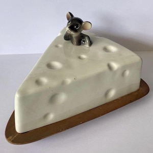 May include: A triangular ceramic cheese-shaped butter dish with a glossy off-white finish and textured indentations. A small, grey and white ceramic mouse figurine is attached to the top lid. The dish rests on a flat, brown wooden base.
