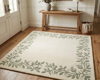 May include: A rectangular area rug featuring a cream-colored base with a sage green botanical border. The border consists of raised, tufted vines, leaves, and small flower blossoms that frame the central plain field of the rug.