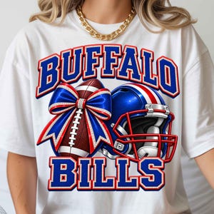 Puede incluir: Una camiseta blanca con un estampado gráfico que muestra el texto BUFFALO BILLS en letras azules con bordes rojos y blancos. El diseño incluye un balón de fútbol americano marrón con un lazo azul y rojo y un casco de fútbol azul con una franja roja.