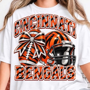 May include: A white t-shirt featuring a graphic with the text CINCINNATI BENGALS in orange and black tiger-striped block letters. The design includes a football adorned with a large orange and black bow and a football helmet with a tiger stripe pattern.