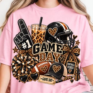 May include: A light pink t-shirt featuring a graphic print of a black foam finger with #1, a leopard print iced coffee, a football helmet, a pom-pom, a football, a baseball cap, sunglasses, and a tag with the text GAME DAY and FOOTBALL VIBES.
